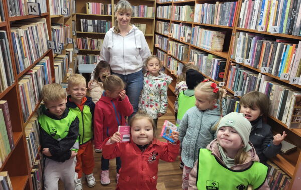 Grupa małych dzieci wraz z bibliotekarką stoi w bibliotece między regałami z książkami. Niektóre dzieci trzymają książki, część ma odblaskowe kamizelki