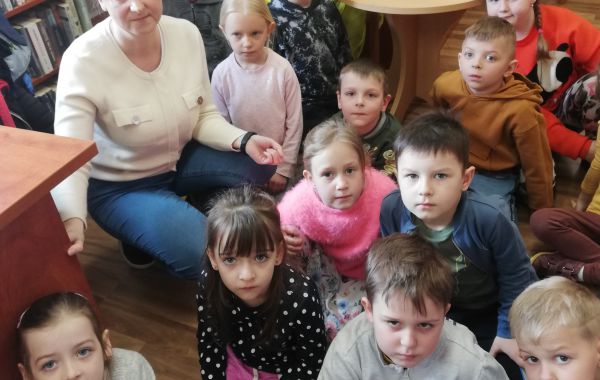 Dzieci siedzące na podłodze. Wśród nich – kucający bibliotekarz