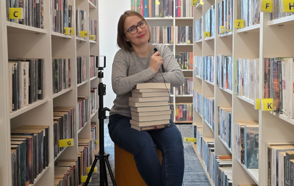 Między regałami z książkami siedzi kobieta z mikrofonem w ręku