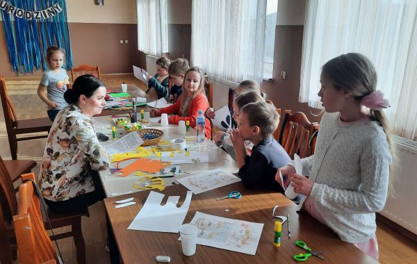 Dzieci siedzące po jednej stronie stołu i wykonujące ozdoby kojarzone ze Świętami Wielkanocnymi.  Po drugiej stronie stołu - bibliotekarz