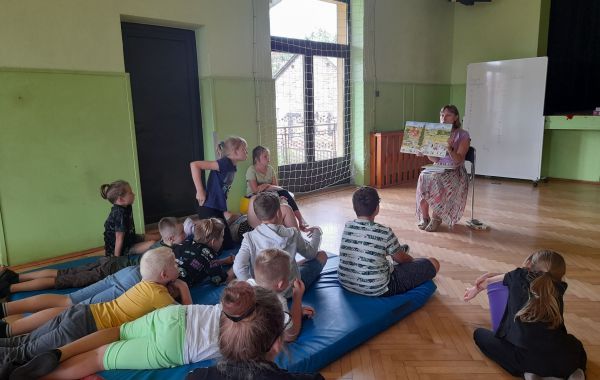 Grupa dzieci siedząca na materacu podczas słuchania czytanej przez bibliotekarza książki