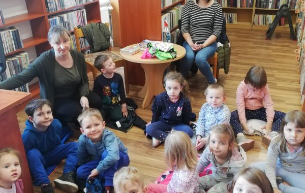 Dzieci siedzące na podłodze przed regałami. Wśród nich – kucający bibliotekarz